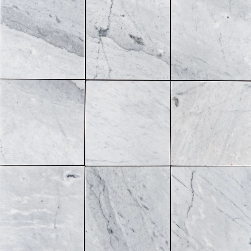 Top-down view of multiple Carrara 6"x6" honed marble tiles with soft white surface and natural gray veining in a grid layout.